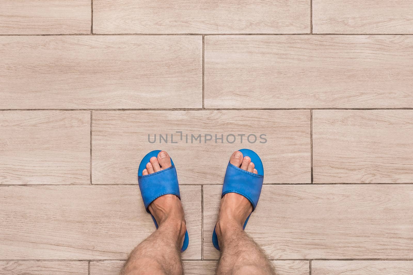 Men's bare feet in blue home slippers stand on a light laminate floor tile background, view from above.