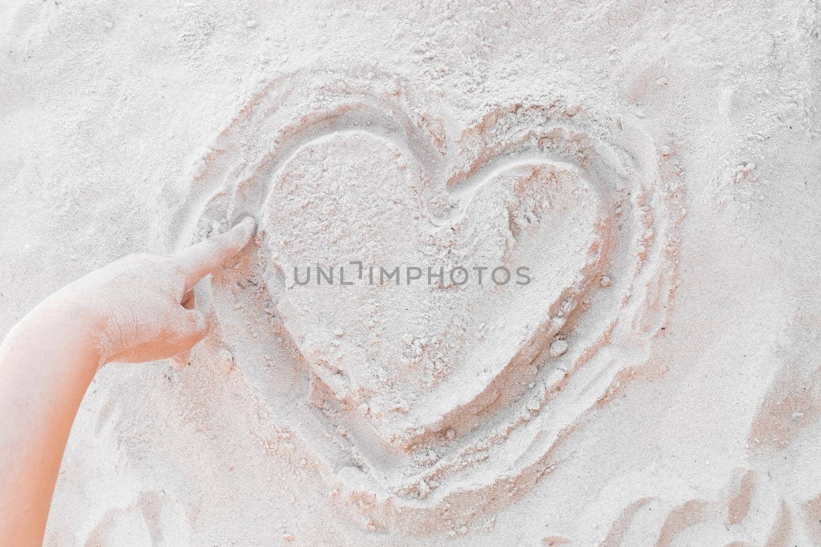 The hand of a young girl draws a heart on the white beach sand close-up. Symbol or sign of love concept.