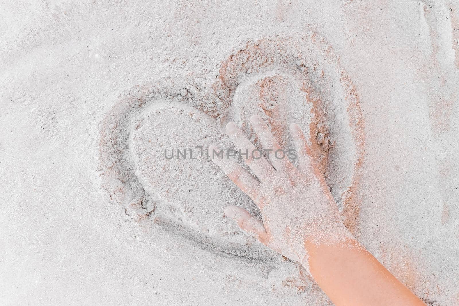 The hand of a young girl touches a picture of a heart on a white beach sand close-up. Sign and symbol of love and relationship concept.