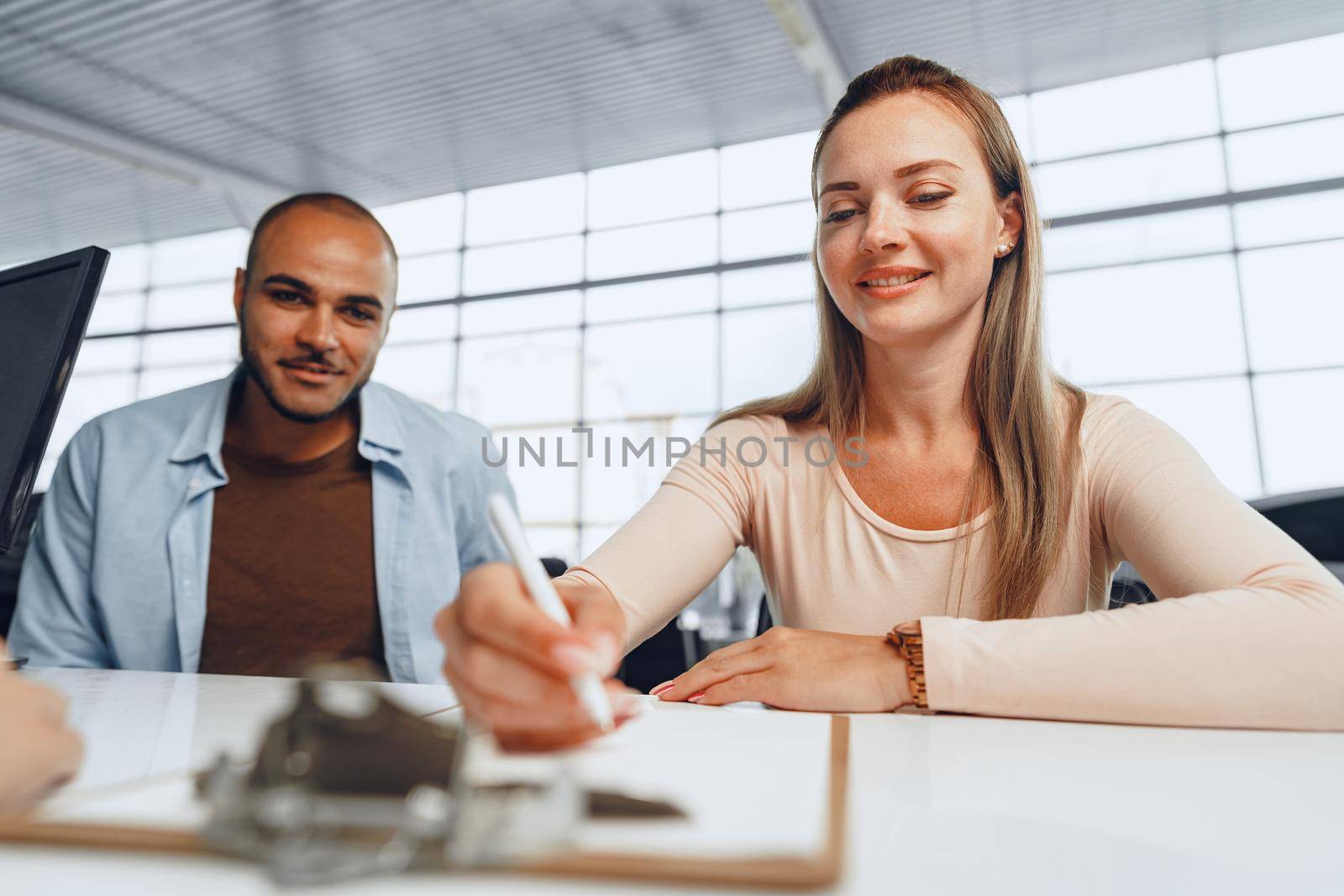 Beautiful young couple signs documents at car dealership showroom sitting at the table