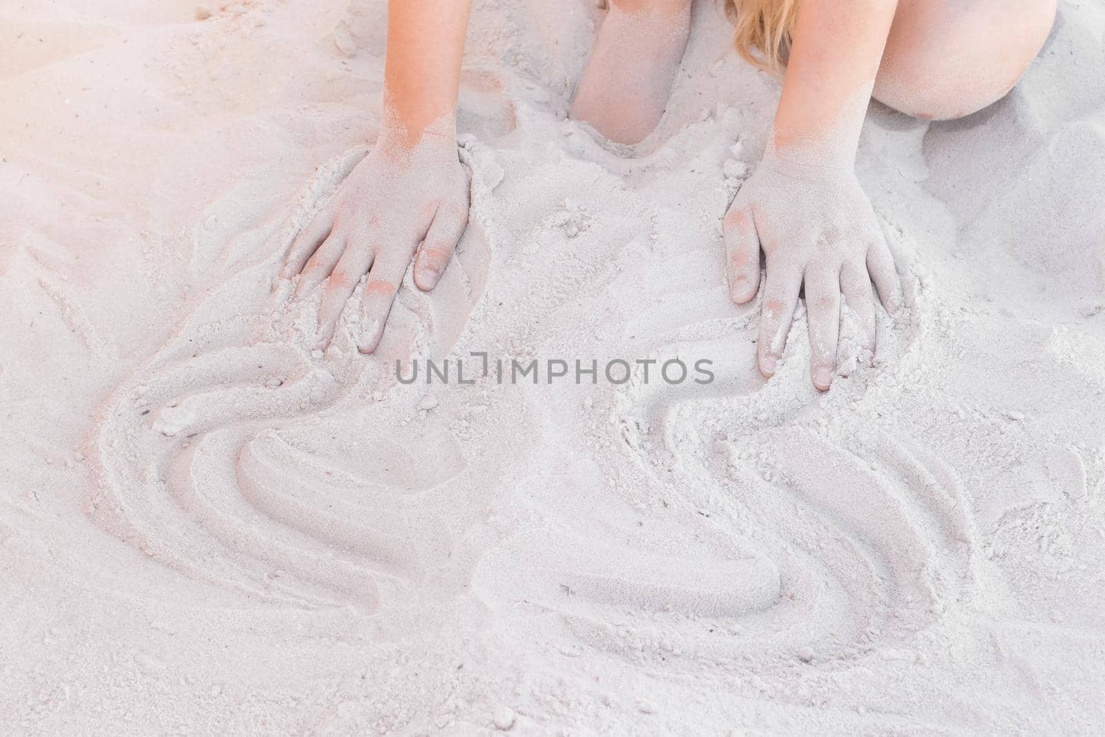 The hands of a young girl draw abstract patterns on a white beach sand background.