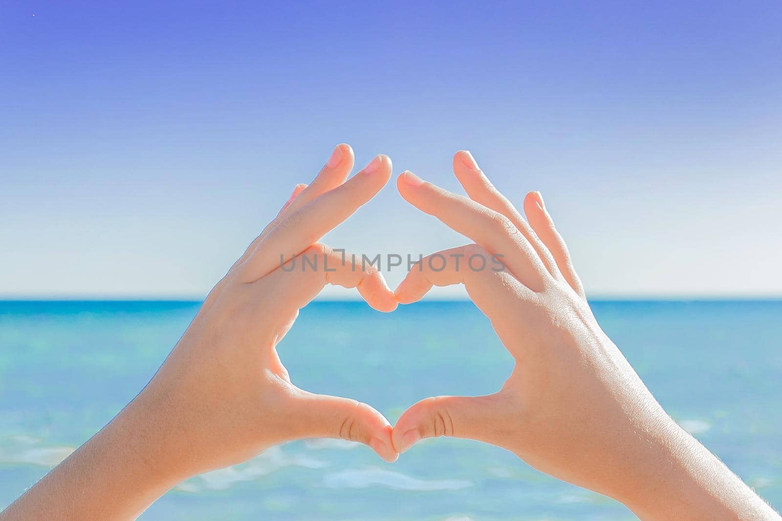 The hands of a young girl depict a sign or symbol of heart and love against the backdrop of the sea and the sky by AYDO8