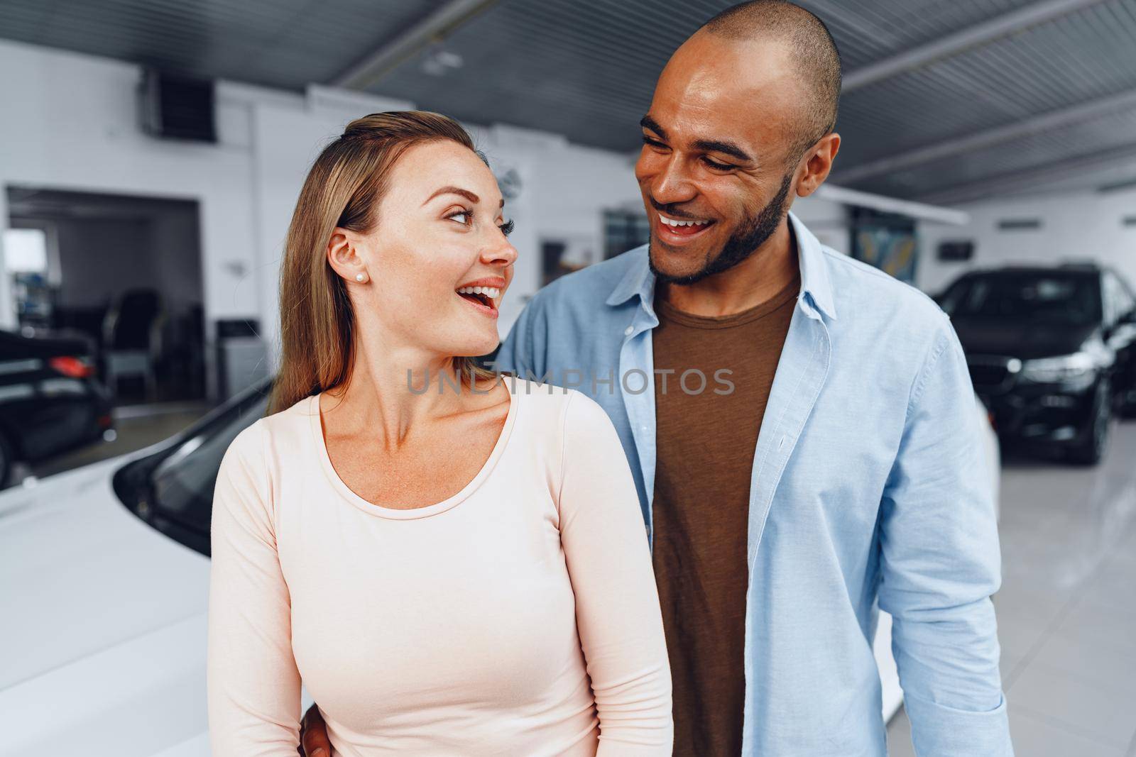 Happy couple of caucasian woman and african american man standing near their new luxury car inside car salon