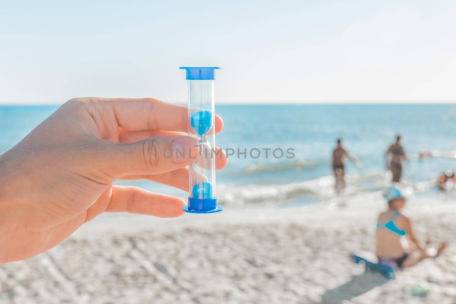 The young man's hand holds an hourglass close-up against the sea beach with holidaymakers in the resort area, blue sea and sky with a skyline by AYDO8