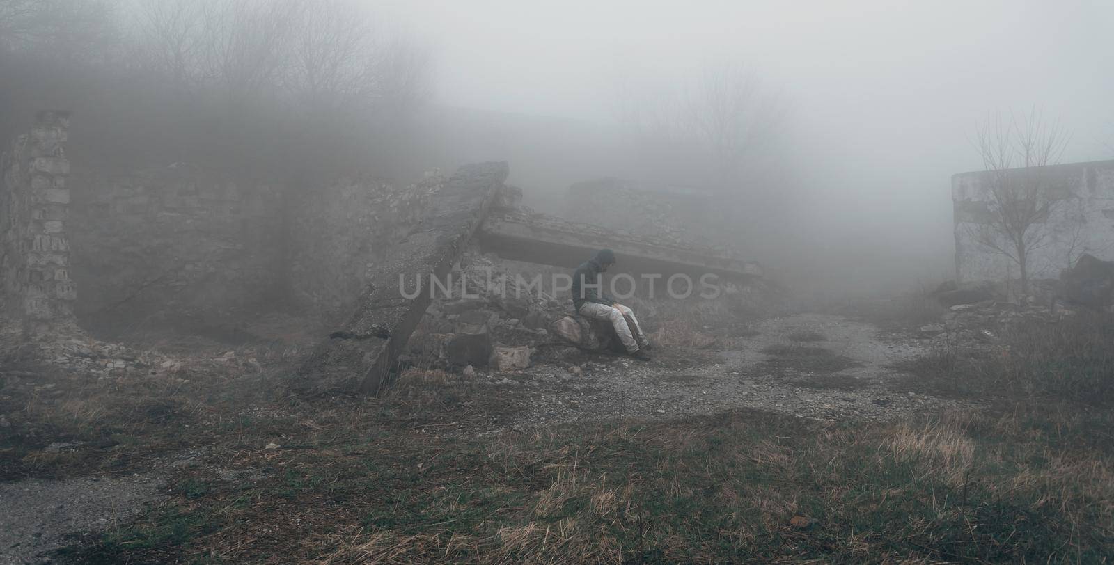 Man in a hood sitting among ruins at mystical mist, post-apocalyptic scene