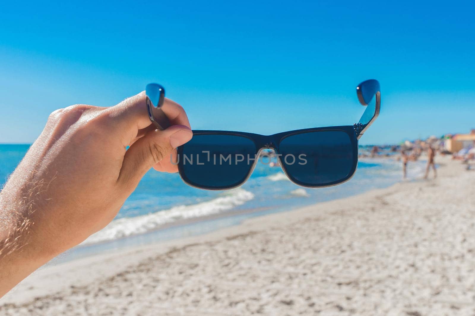 A young man's hand holds sunglasses against a sea beach by AYDO8