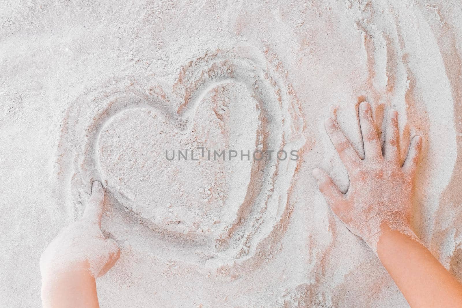 The hand of a young girl draws a heart on the white beach sand close-up. Symbol or sign of love concept.