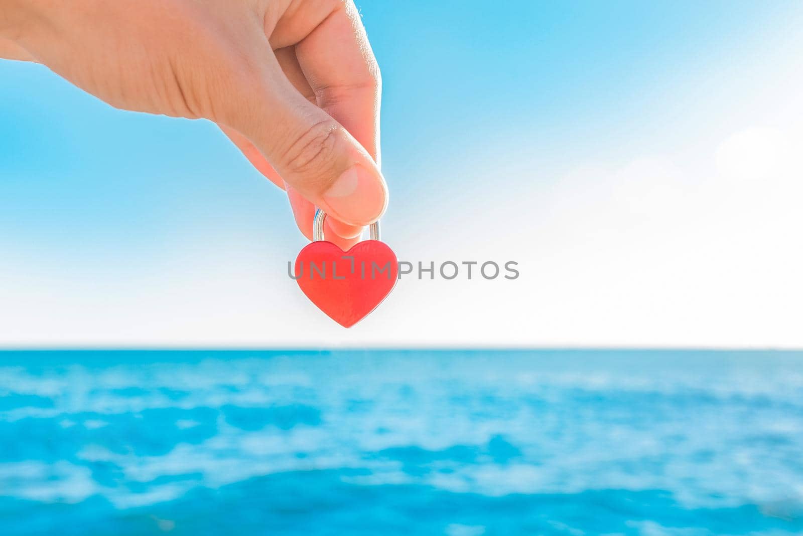 The hand of man holds a red little castle in the form of a heart close-up against the blue sea and the sky with a horizon line. Symbol or sign of love.