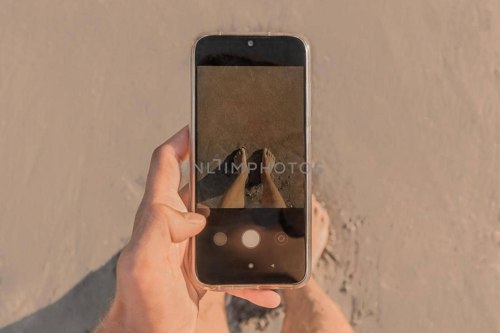 The man's hand holds a mobile phone and takes pictures of his bare feet standing on the wet beach sand by the sea, close up.