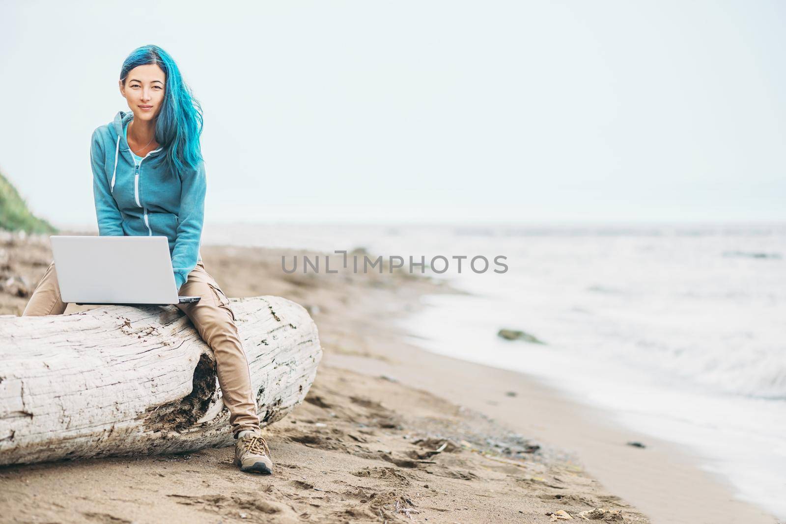Smiling freelancer girl with blue hair working on laptop on sand beach near the sea, looking at camera. Freelance concept