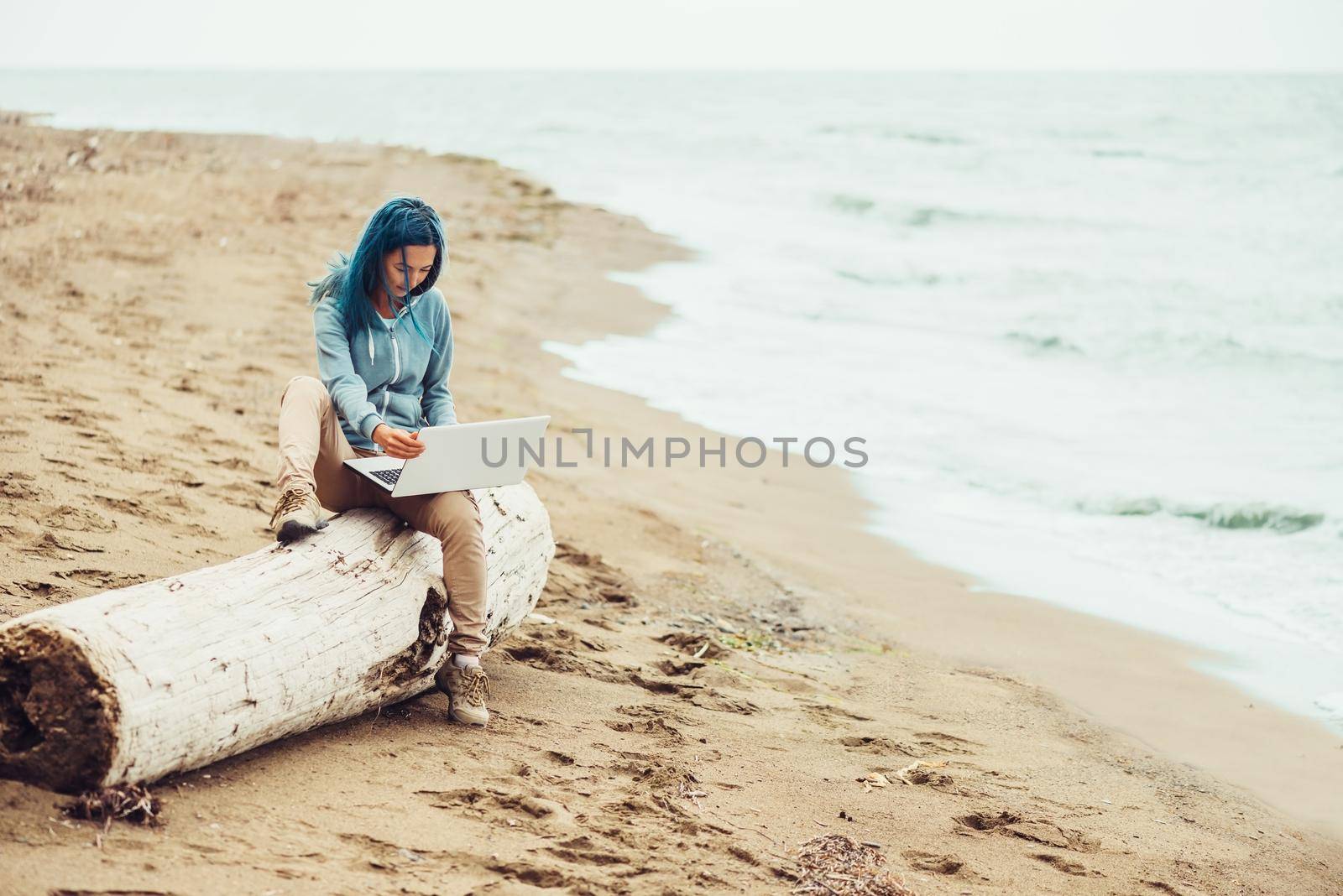 Freelancer young woman with blue hair sitting on tree trunk and working on laptop on sand beach near the sea. Freelance concept