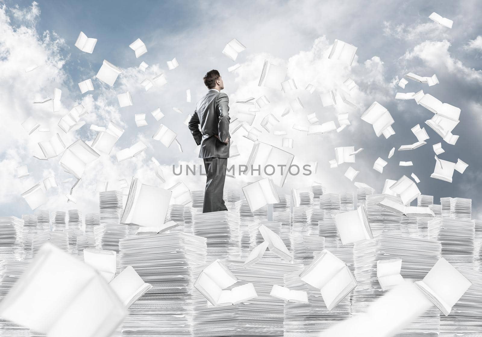 Confident businessman in suit standing on pile of documents among flying books with cloudly sky on background. Mixed media.
