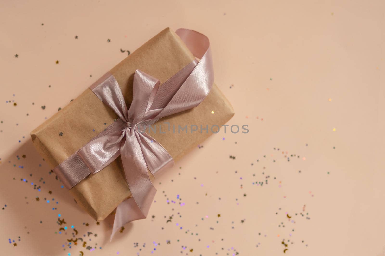 Birthday or New Year's eve present in kraft paper with pink ribbon on a soft beige background with glitter and confetti. Xmas composition. Flat lay. Happy holidays celebration and giving love concept