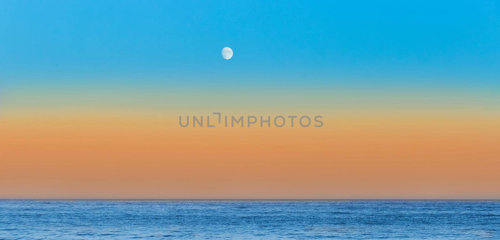 Twilight on the sea beach in the evening with an orange sunset on skyline and blue sky with a moon.