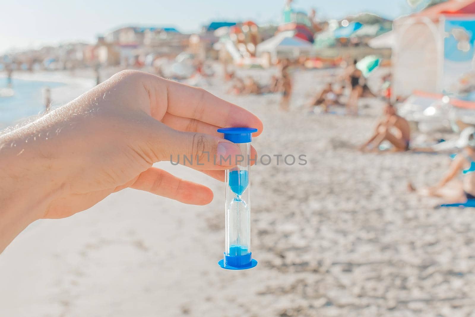 The guy's hand holds an hourglass against the backdrop of a sea beach with holidaymakers in the resort area, close up.