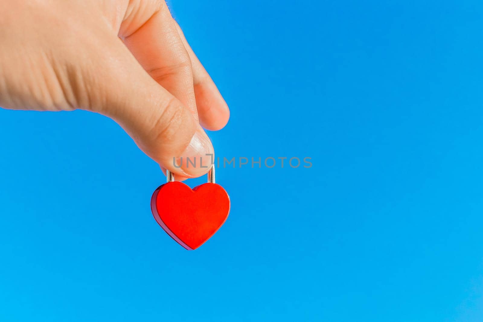 The hand of a man holds a small red heart in the form of a castle close-up against the blue sky. Symbol and sign of love by AYDO8