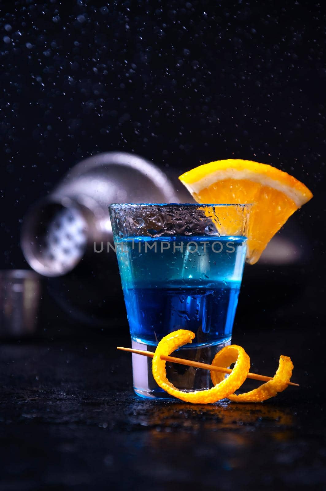 Shot with White Rum, Liquor Blue Curacao and Orange Slice. Alcoholic Layer Cocktail with Shaker in Freeze Motion, Drops in Liquid Splash on Dark Background.