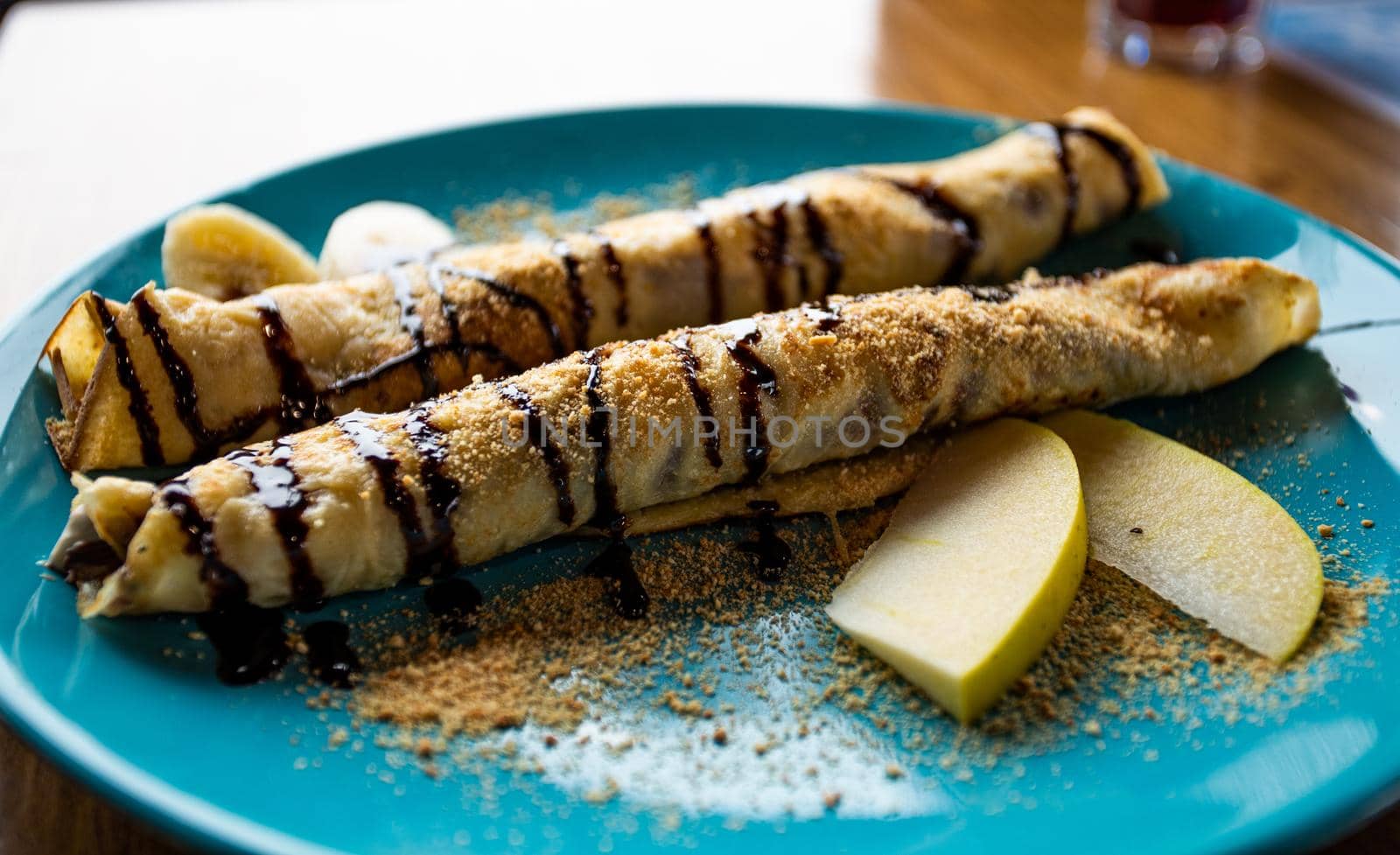 Arranged two crepe pancakes rolls on blue plate with apple and banana slices and chocolate topping. Dessert or sweets on table in restaurant