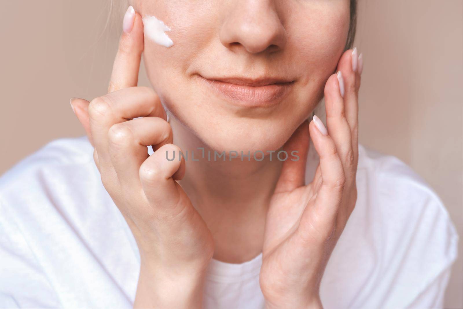 Beautiful smiling girl applying cream on face. Body, skincare concept. Woman in white with natural pink manicure uses beauty products. Skin protection. Moisturizing lotion. Young female closeup