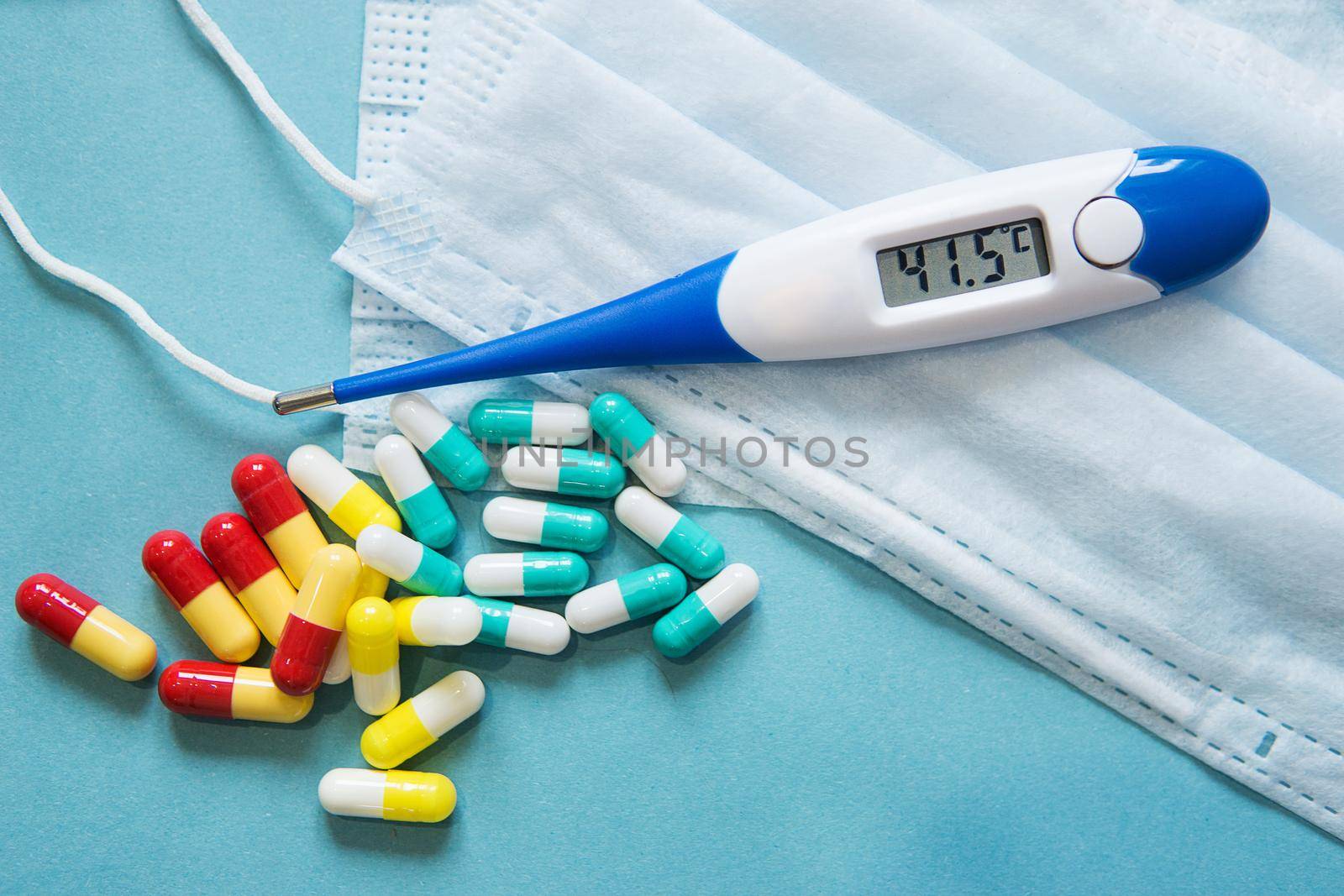 Respiratory mask, thermometer with high temperatureand medicine in capsules on blue background.Virus prevention and treatment.