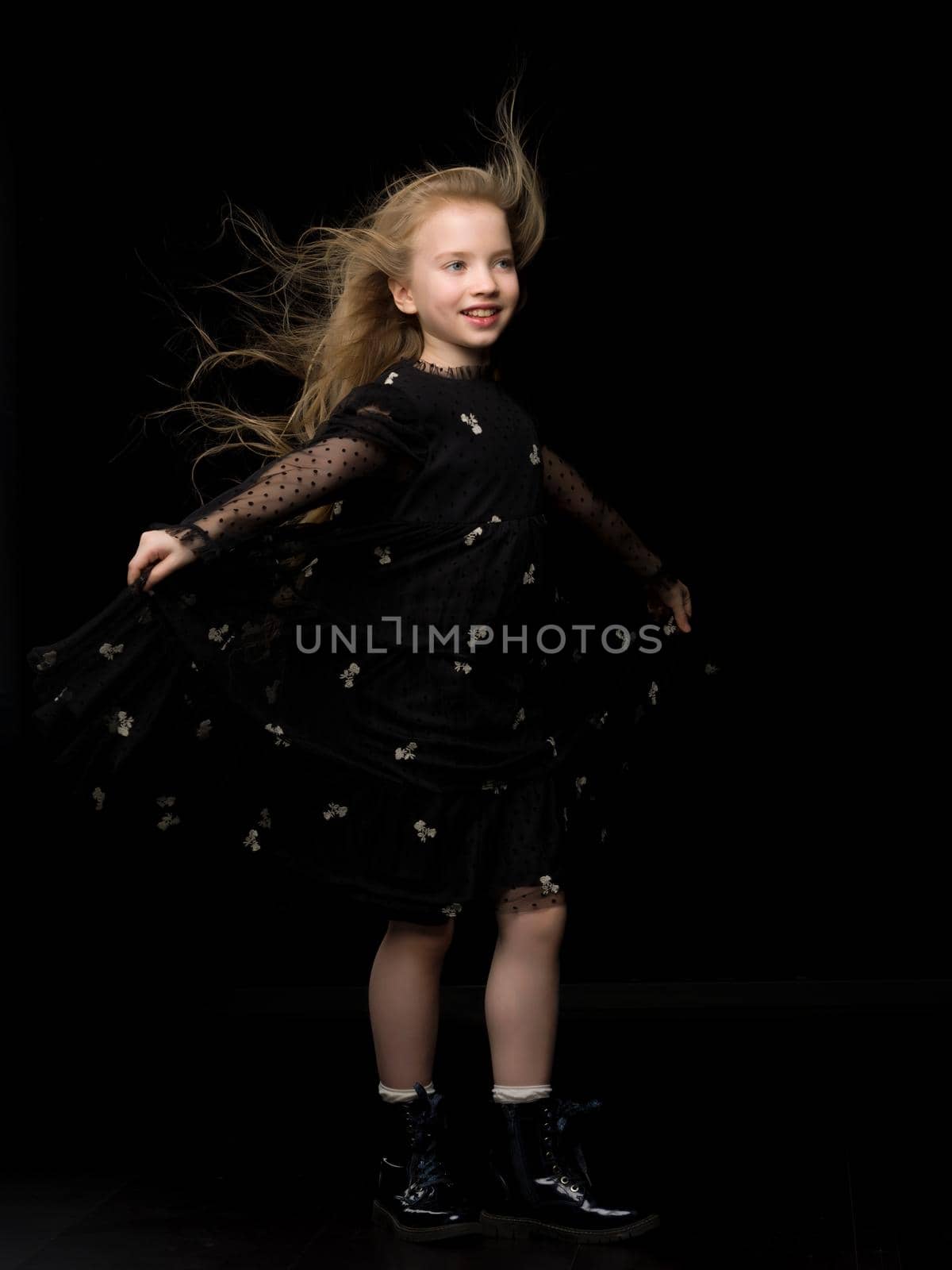 A strong wind blows on the face and clothes of a beautiful little girl of school age. Hair and clothes are fluttering in the wind, and the girl is happy about it. On a black background. Studio photo session