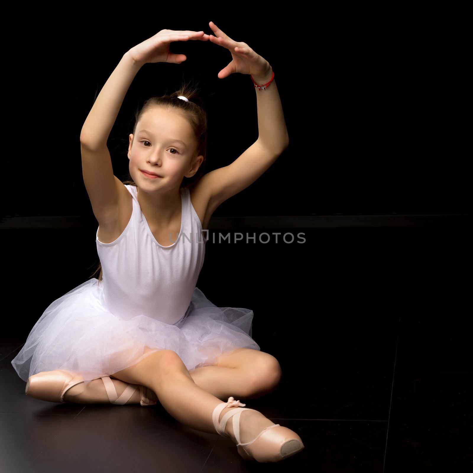 Adorable little girl ballerina. She is posing in pointe shoes, sitting on the floor in the studio. Isolated on a black background.