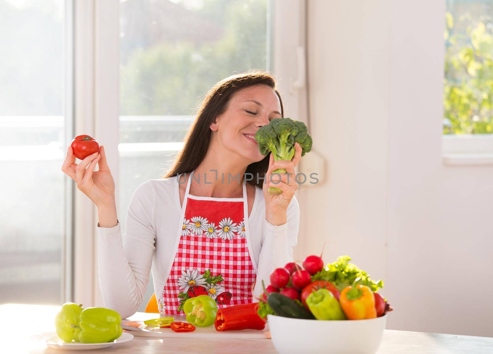 Satisfied woman sitting at kitchen table with vegetables in bowl and plates and enjoying smell of broccoli