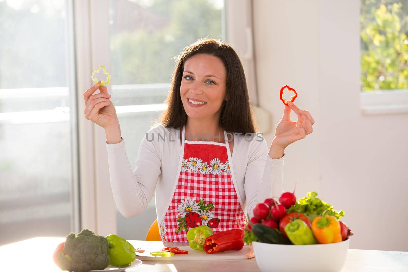 Happy young woman sitting at table with vegetables in bowl and holding pepper slices in hands