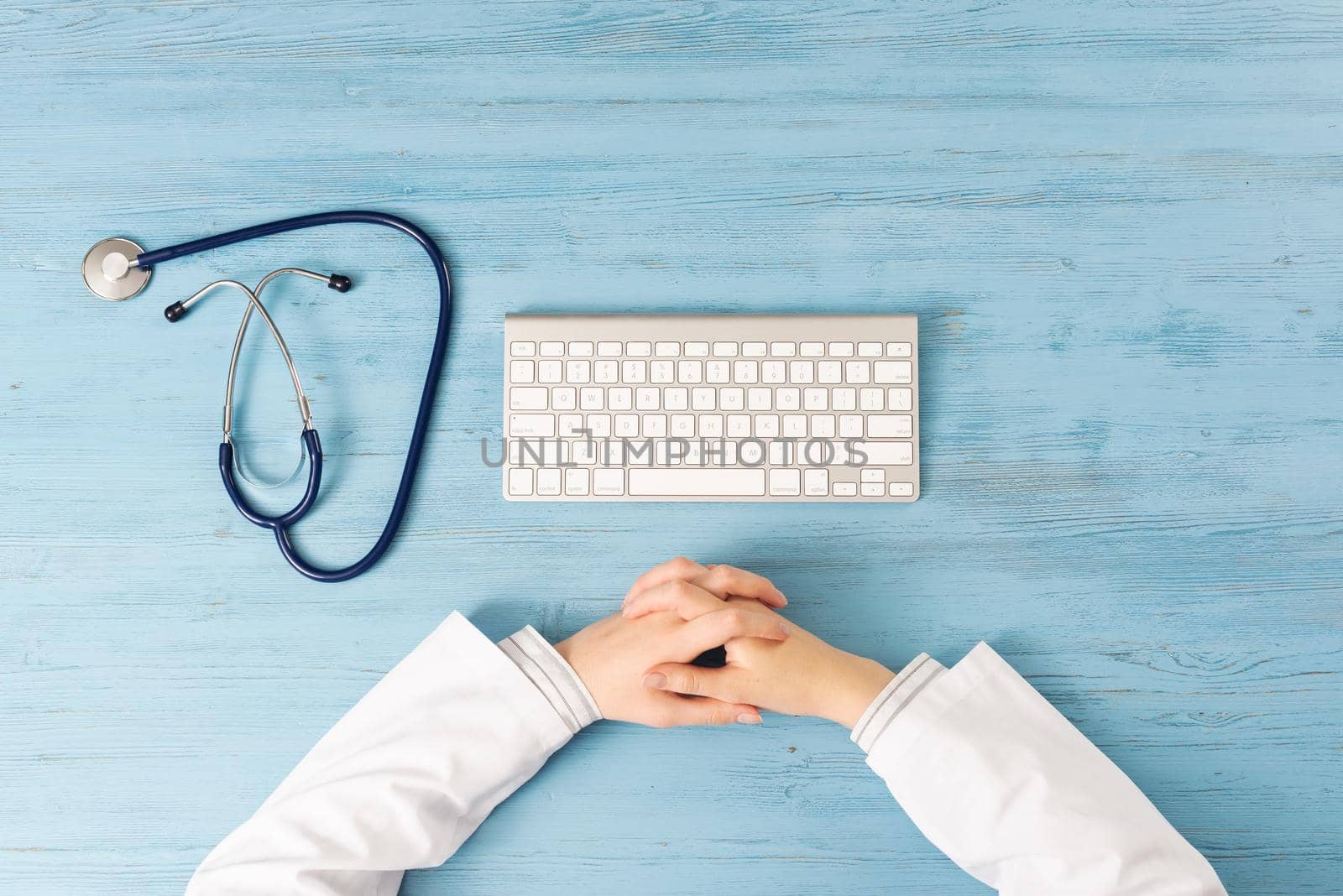 Top view of doctor with folding hands. Physician in medical uniform working at blue wooden desk. Examination and diagnosis in hospital. Practitioner workplace with stethoscope and computer keyboard.