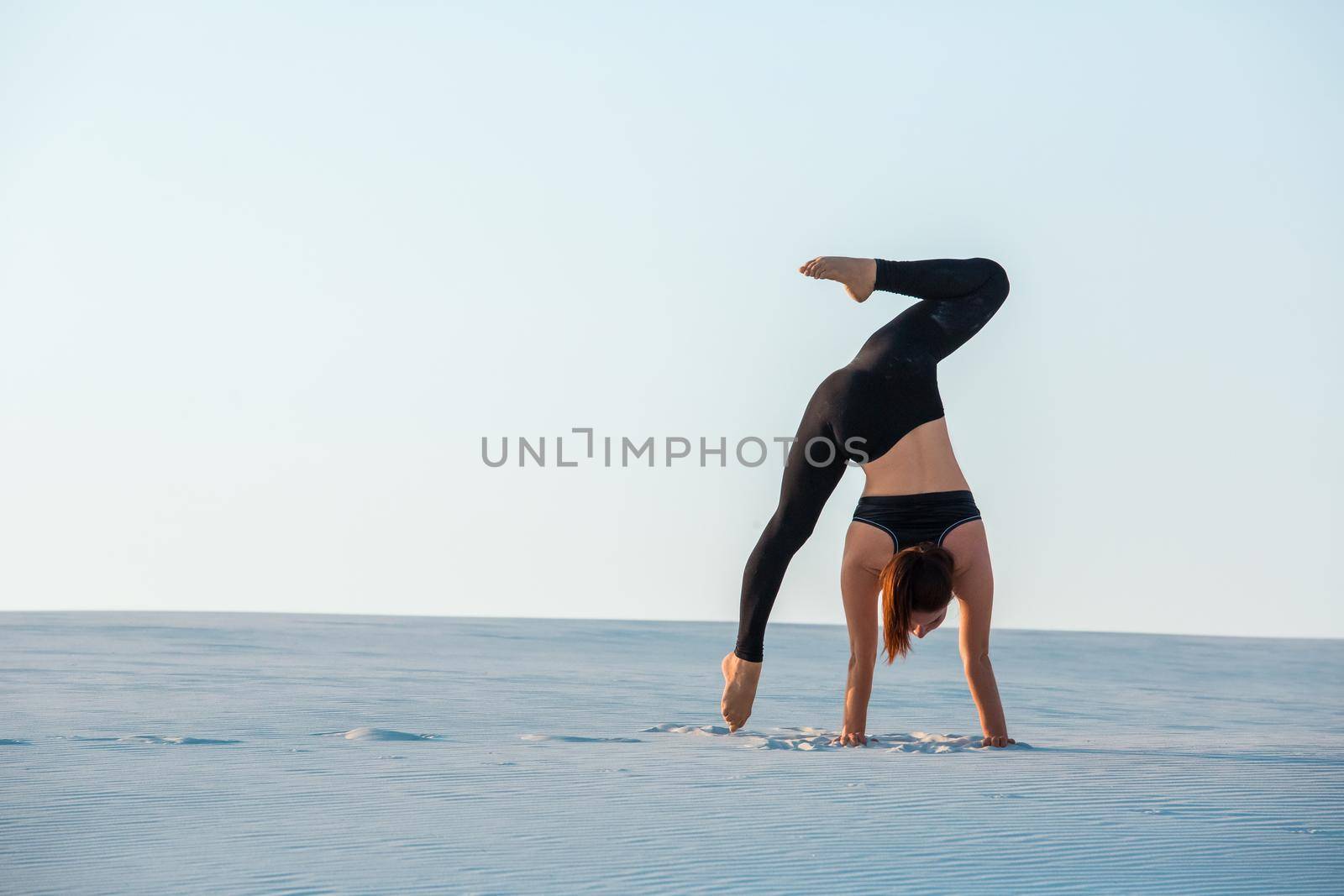 Young woman practicing inversion balancing yoga pose handstand on sand. Sunset and white sand.