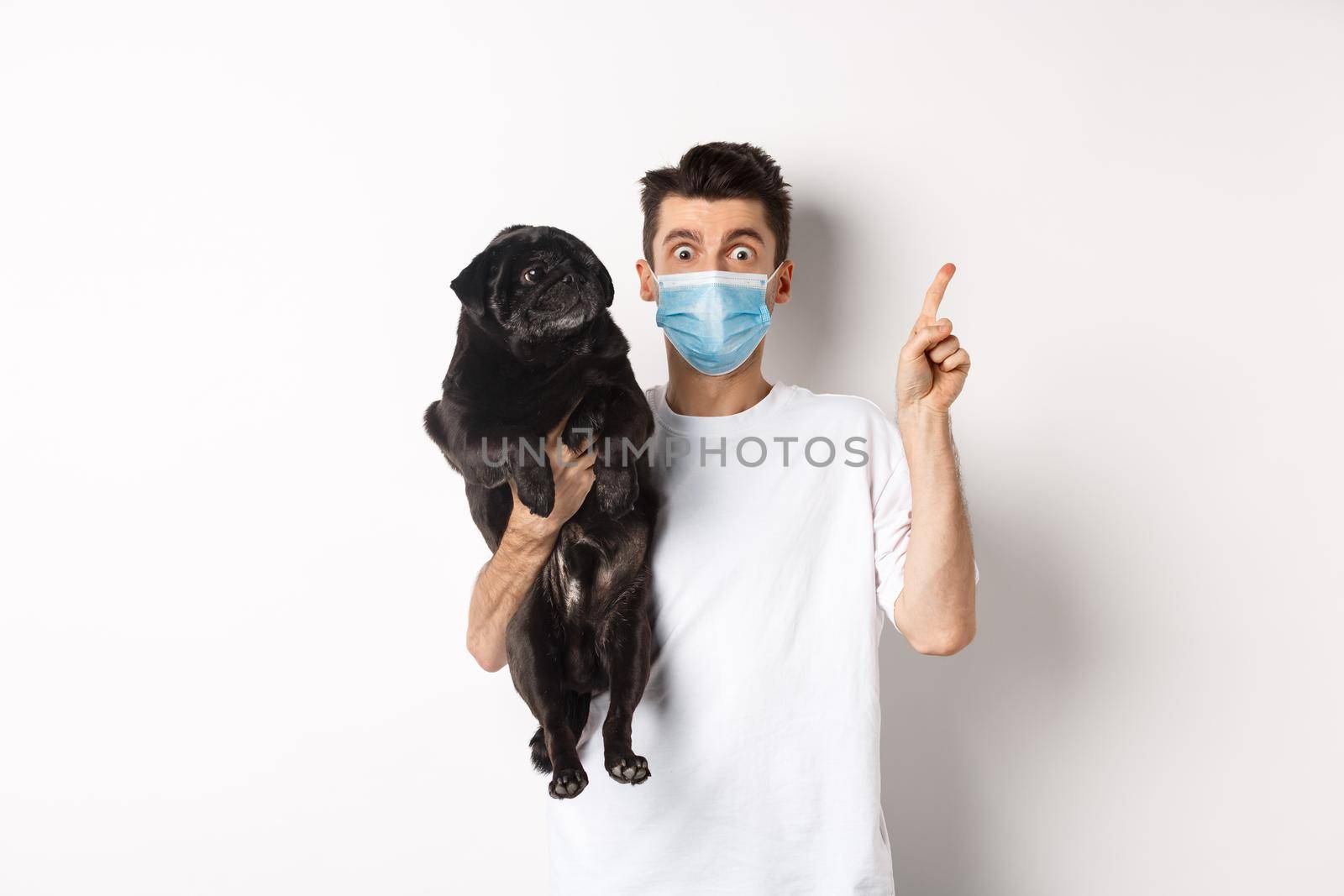 Covid-19, animals and quarantine concept. Young man in medical mask holding cute black pug, dog looking right and owner pointing at logo, white background by Benzoix