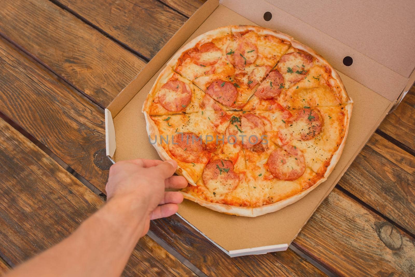 The guy's hand takes a pizza in cardboard box of against the background of a wooden table. Delicious fast food by AYDO8