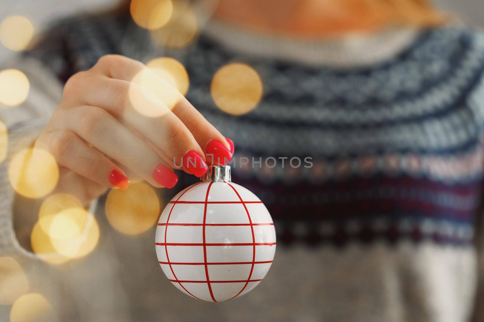 Hand of a woman in festive sweater holding Christmas bauble with blurred lights close up