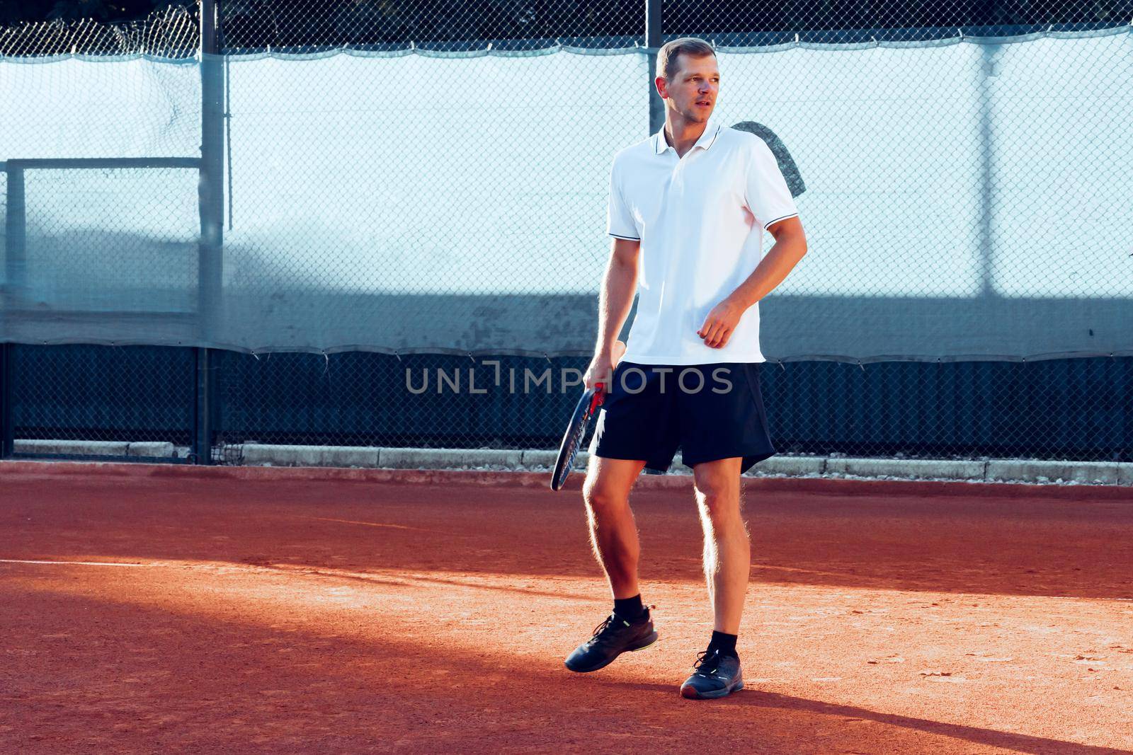 Young man plays tennis outdoors on clay tennis court in the morning
