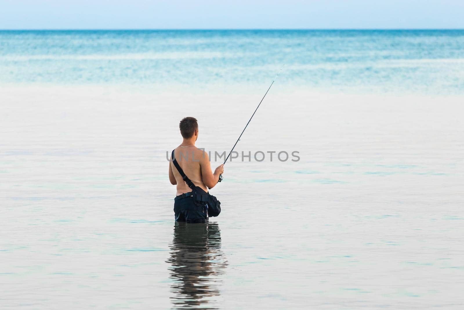 A young guy without a shirt stands knee-deep in the water of the sea in the evening and catches fish with a line by AYDO8