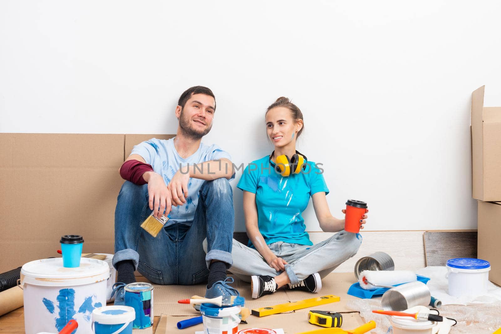 Happy boy and girl drinking coffee on floor. Home remodeling after moving. Cardboard boxes, construction tools and materials for building on floor. Couple having fun in time of house renovation.