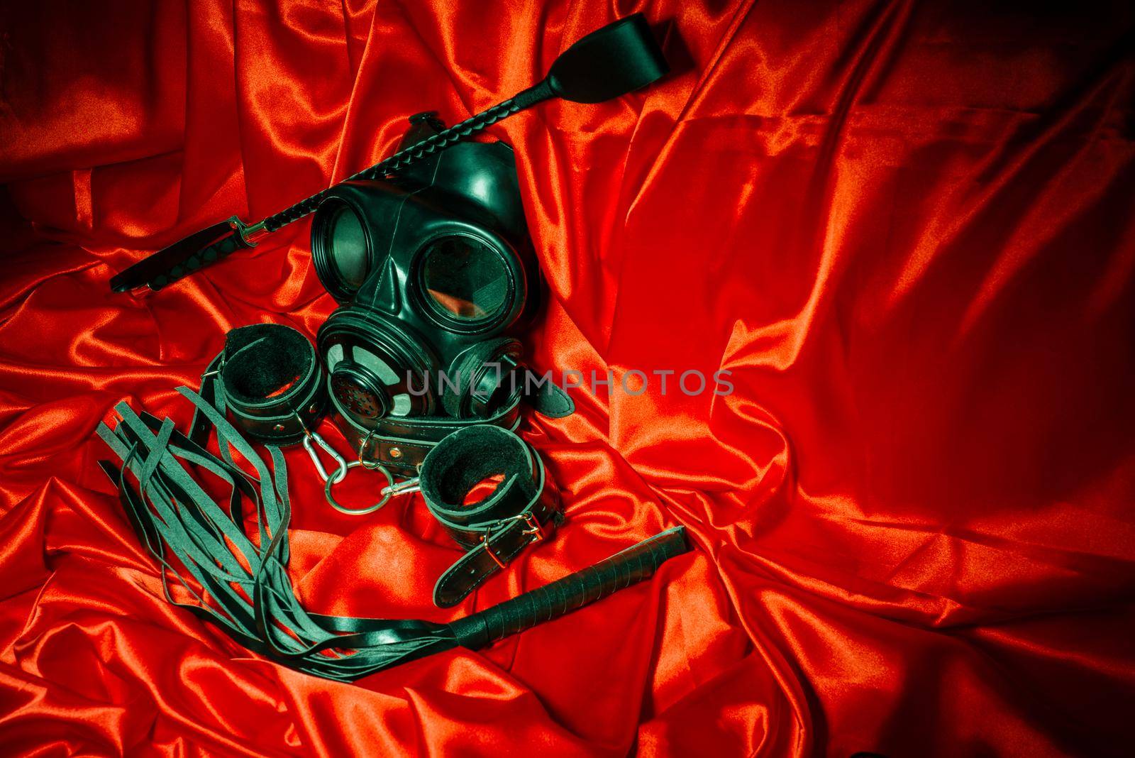 Close up bdsm outfit. Bondage, kinky adult sex games, kink and BDSM lifestyle concept with gas mask, whip, collar, leather handcuffs , lash on red silk with copy space.