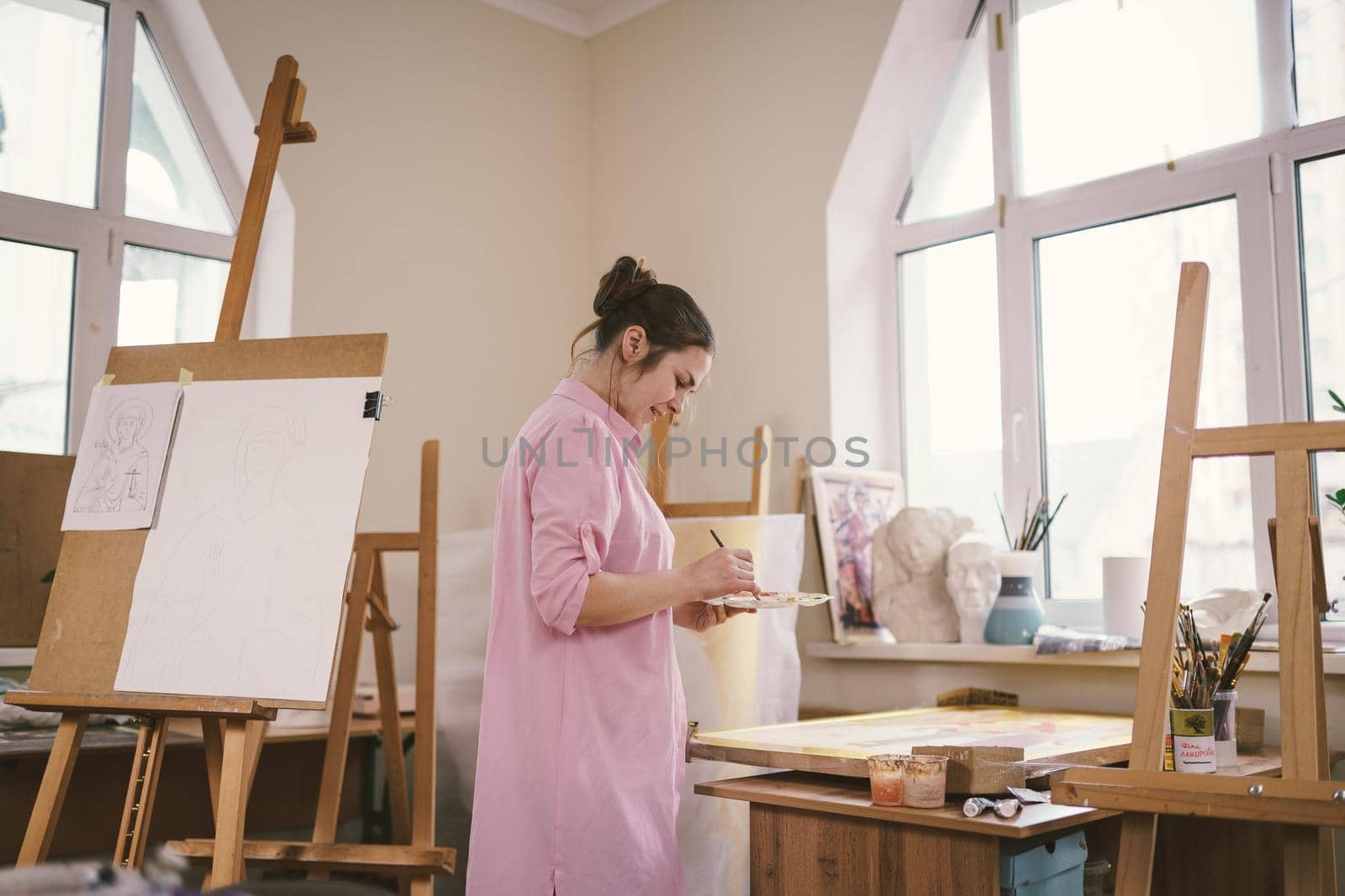 Caucasian woman artist working on a painting in bright daylight studio. Happy artist draws an art project with paints and a brush in the workshop. Hobby. Artist at work. Creative profession by Tomashevska