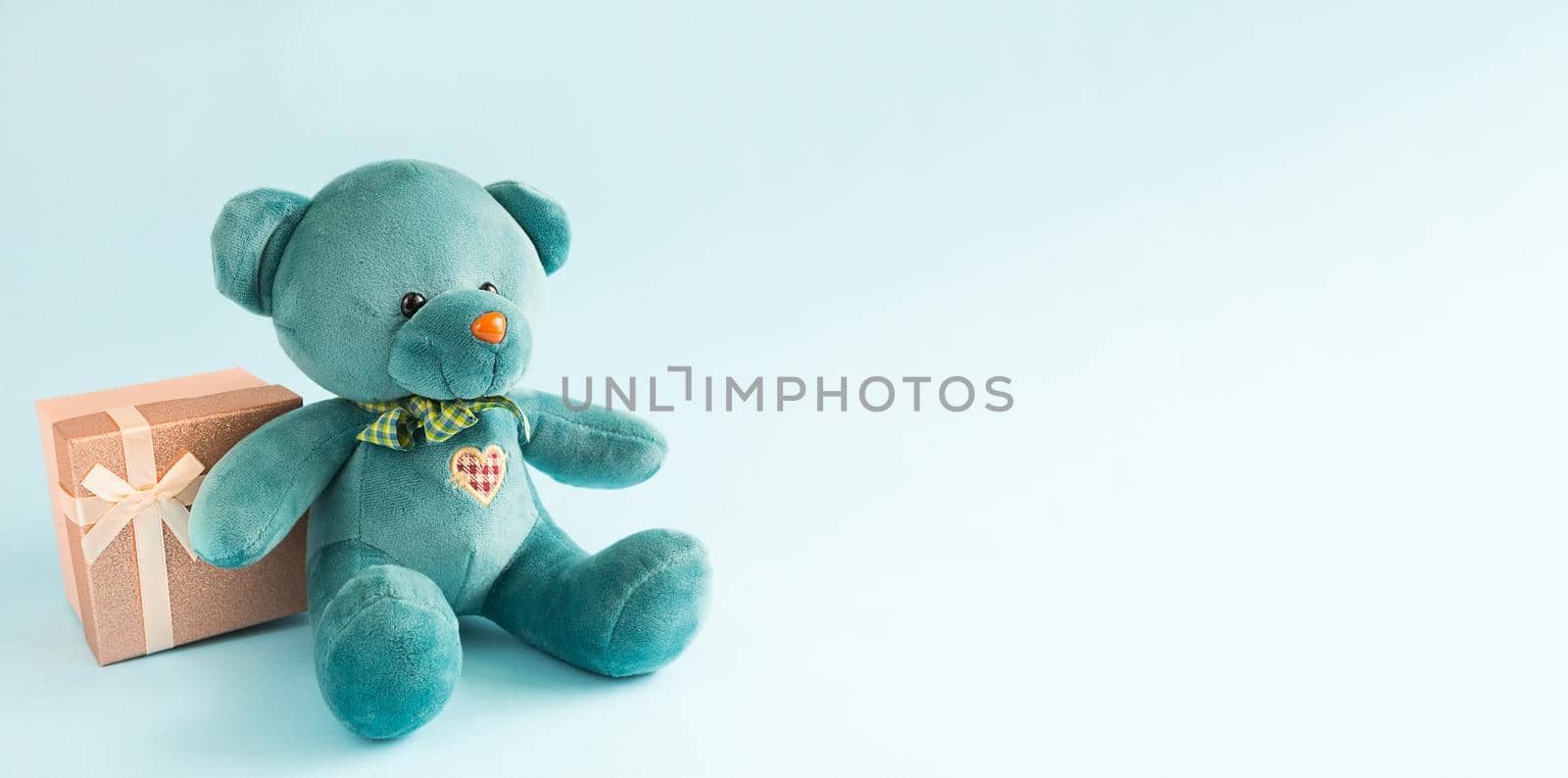 Turquoise soft teddy bear with an embroidered heart holds a gift box and a bow on a blue background. Children's toy. Love, a gift to holiday, a declaration of love, Valentine's day. Copy space by Simol