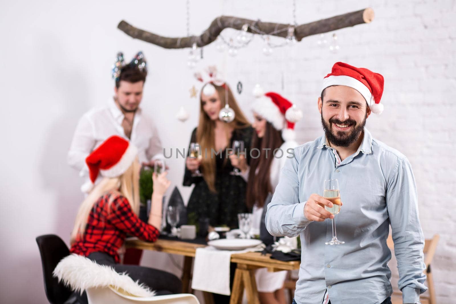 Shot of happy friends enjoying holidays. Focus on the man in the foreground in a red Christmas hat. Man with a glass of champagne in a white room