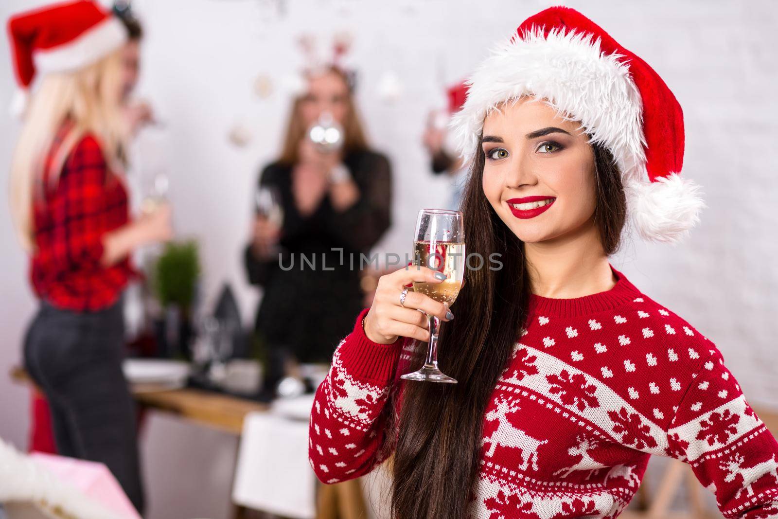 Portrait of a young woman with a glass of champagne at home on the foreground. Beautiful brunette in a Santa hat, red costume with deers by nazarovsergey