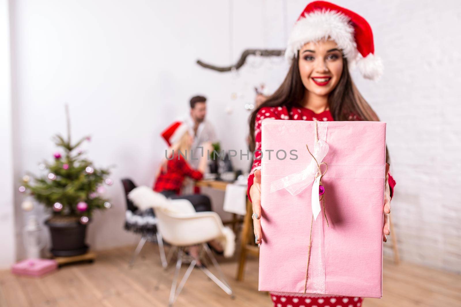 Portrait of a young woman with a pink giftbox on the foreground. Beautiful brunette in a Santa hat, red costume with deers. Christmas party at home.