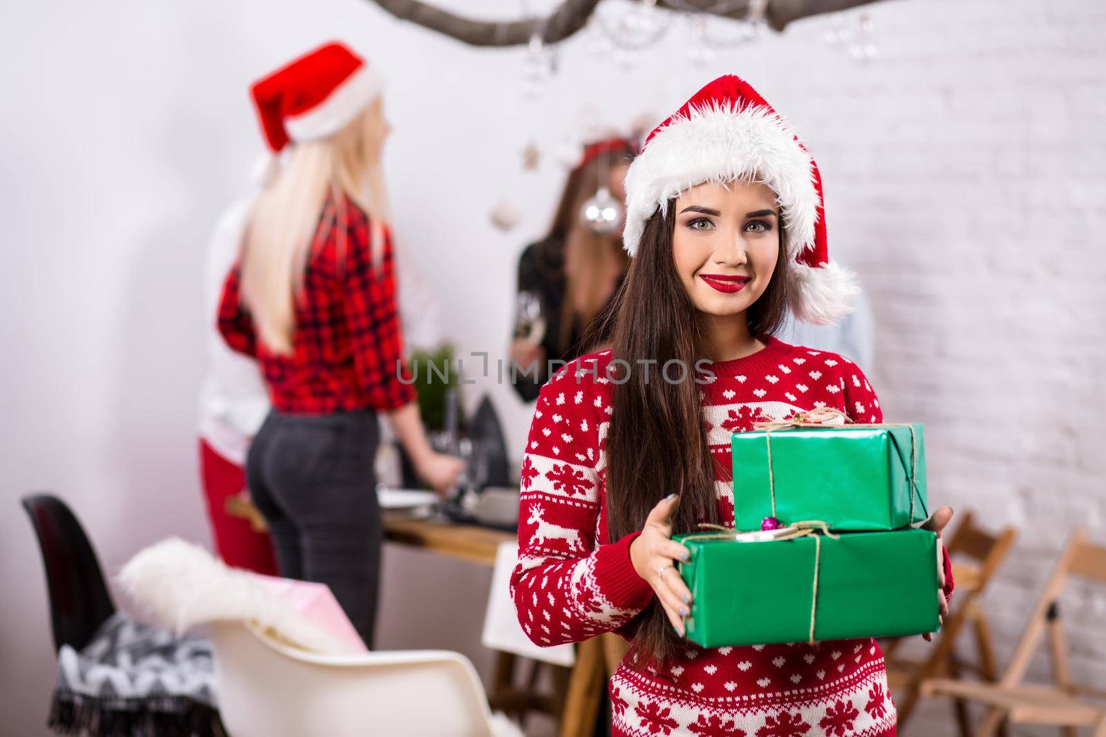 Portrait of a young woman with green giftboxes at home on the foreground. Beautiful brunette in a Santa hat, red costume with deers by nazarovsergey