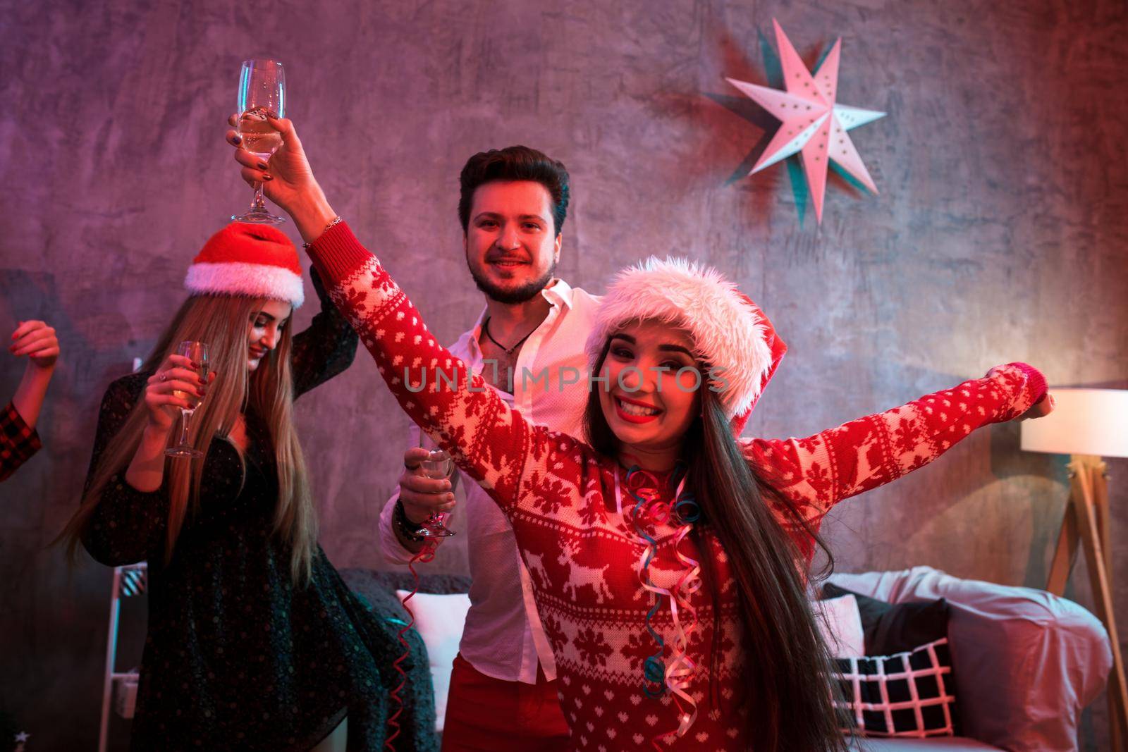 Portrait of a young woman with a glass of champagne on the foreground. Beautiful brunette in a Santa hat, red costume with deers. Christmas party at home.