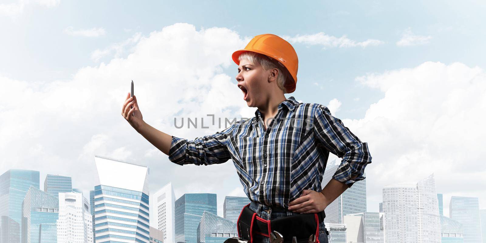 Expressive blonde woman in workwear and hardhat shouting into smartphone. Portrait of young emotional worker in checkered blue shirt standing with open mouth. Business communication and conversation.