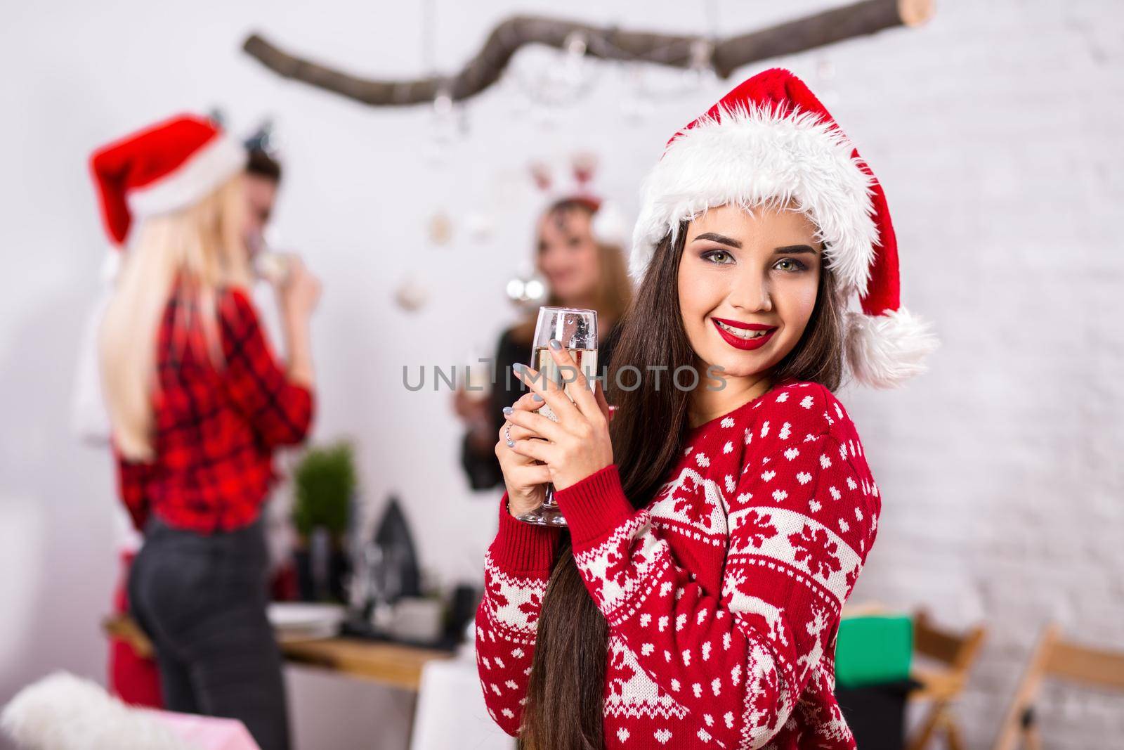 Portrait of a young woman with a glass of champagne at home on the foreground. Beautiful brunette in a Santa hat, red costume with deers by nazarovsergey