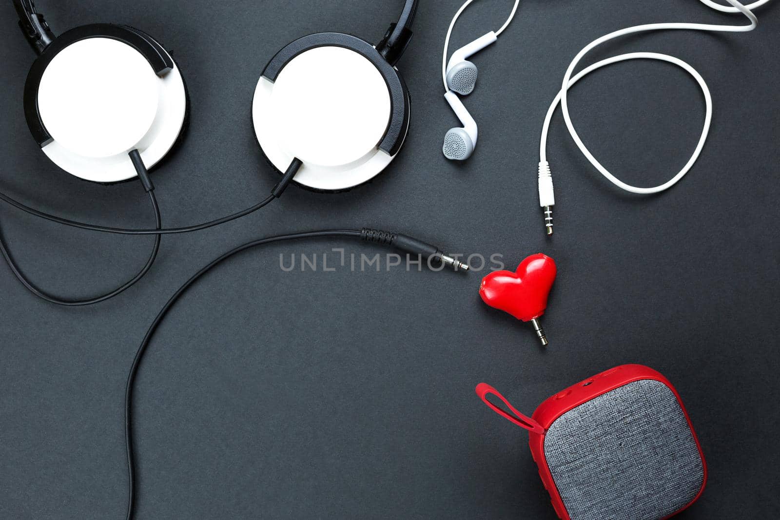 A heart-shaped headphone splitter for a couple in love. Connected to red portable speaker and two pairs of headphones - flat lay. Valentine's day, love, modern technology, copy space, black background by Simol