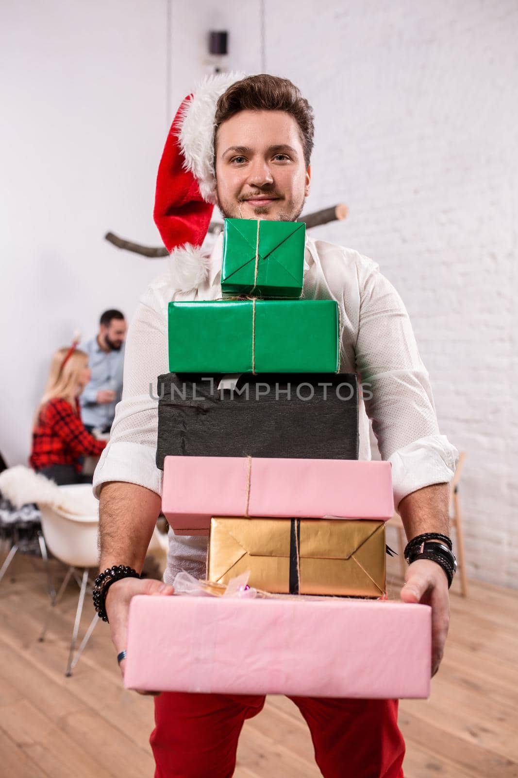 Shot of happy friends enjoying holidays. Focus on the man in the foreground in a red Christmas hat. A man with a gift boxes in his hands in a white room