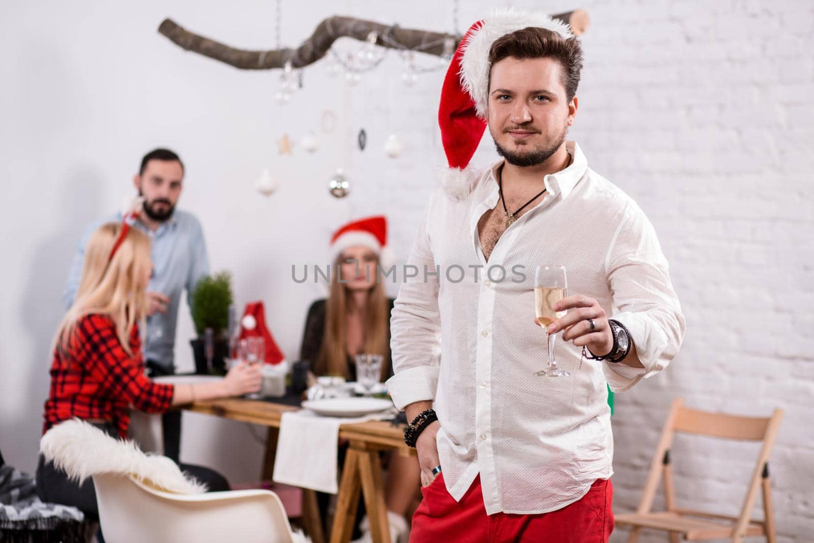 Shot of happy friends enjoying holidays. Focus on the man in the foreground in a red Christmas hat. Man with a glass of champagne in a white room