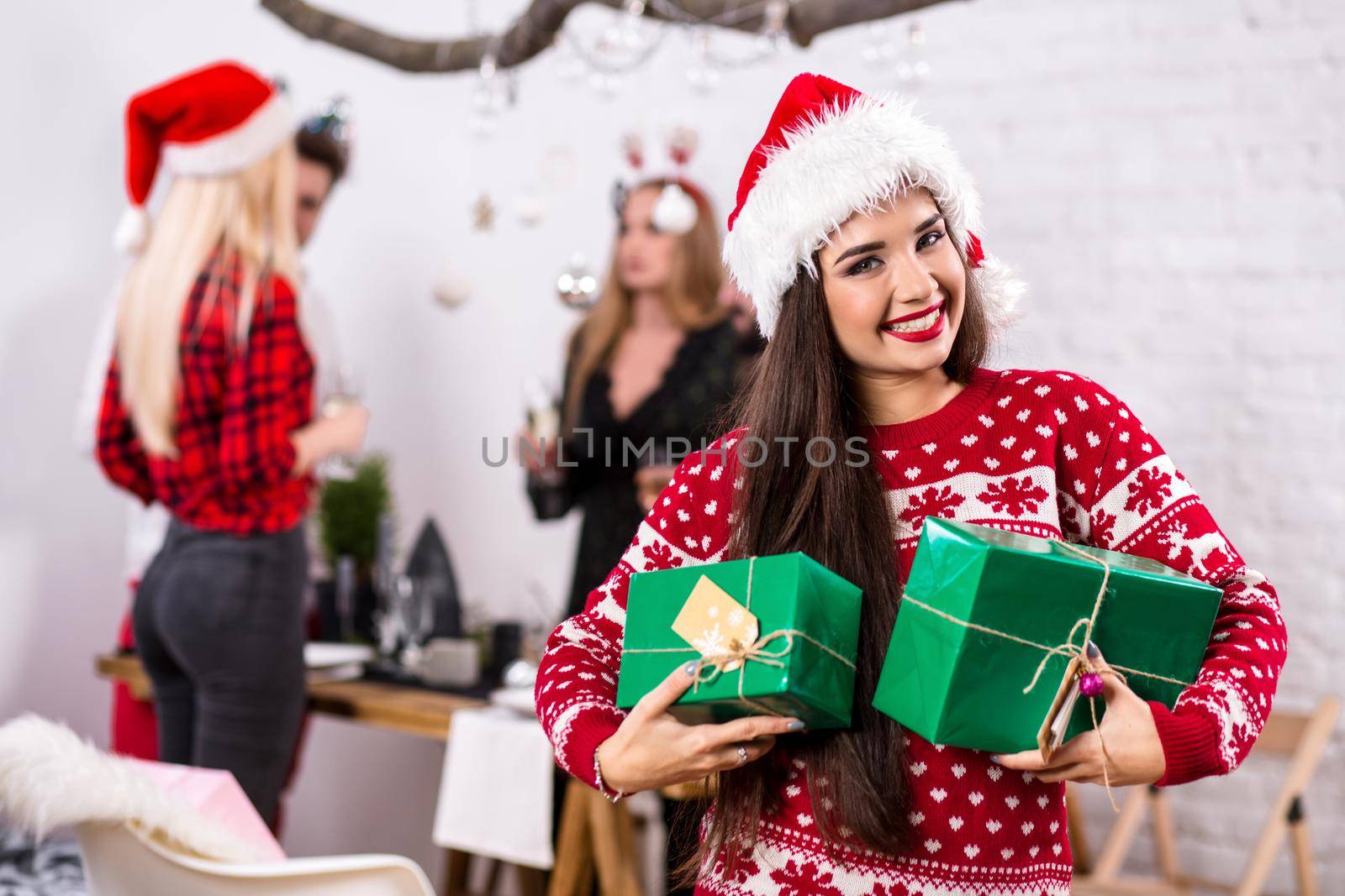 Portrait of a young woman with green giftboxes on the foreground. Beautiful brunette in a Santa hat, red costume with deers. Christmas party at home.